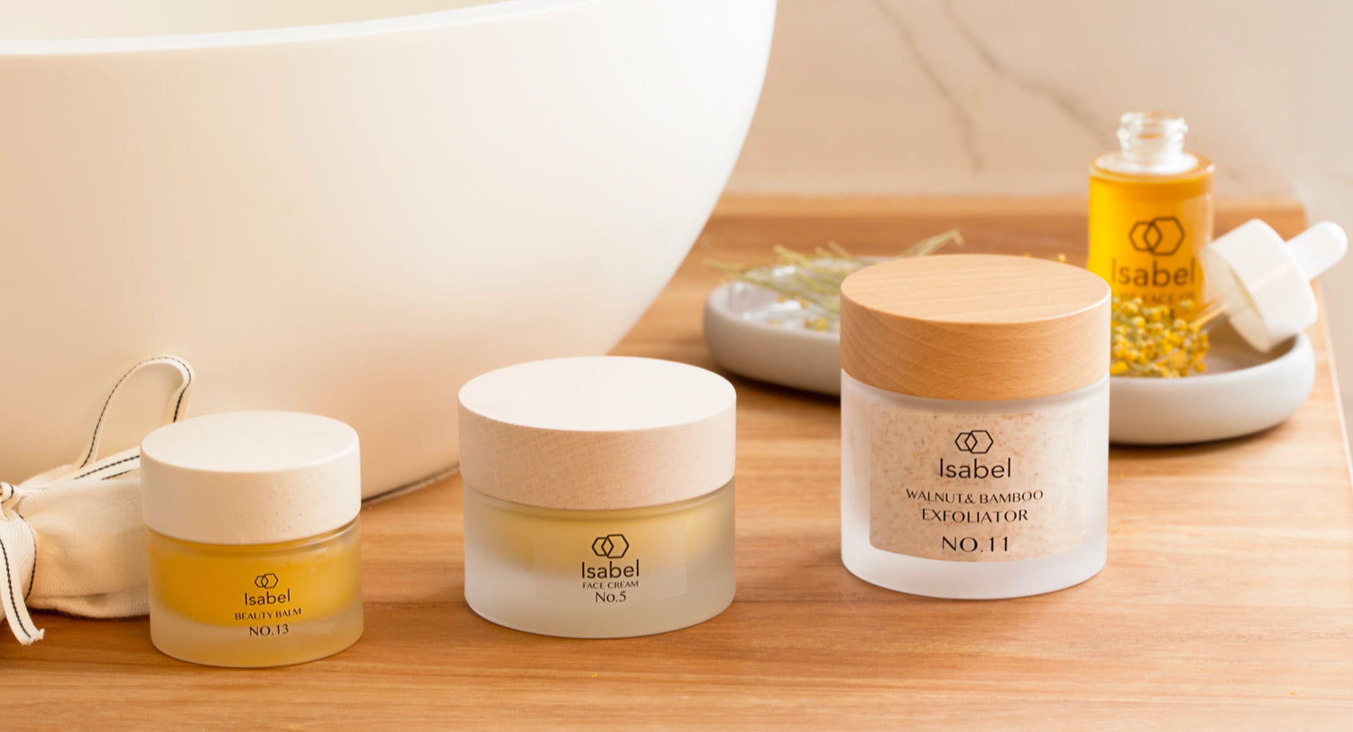 Isabel natural skincare products including face cream, beauty balm, and walnut bamboo exfoliator in eco-friendly jars on a wooden spa surface—luxury clean beauty essentials.